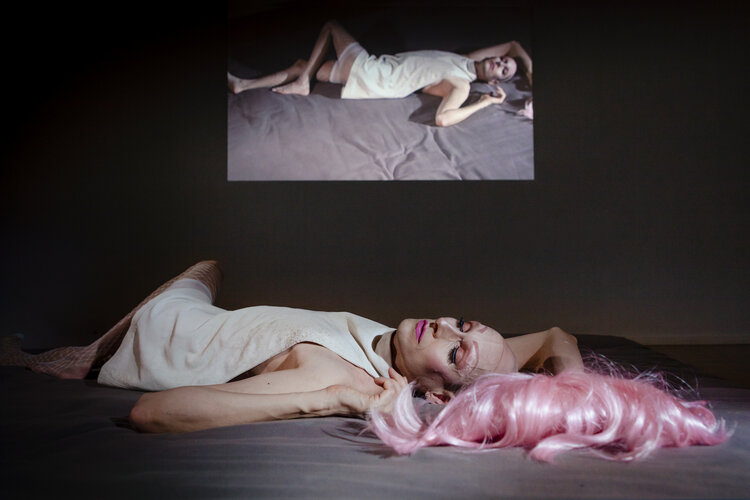 A person is lying on a bed. They have a strong makeup: Pink eyebrows, eye shadow and lipstick. A pink wig has fallen from the head next to the person. They are wearing a white short dress. An image of a person on a bed is projected onto the background.