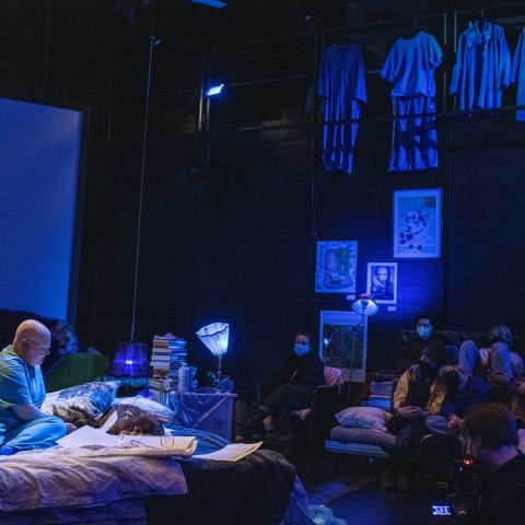 A blue-tinted room with a bald person in blue clothes sitting on a bed on a stage. At the edges of the space there are sofas, pillows and blankets on which people sit. Most have face masks.