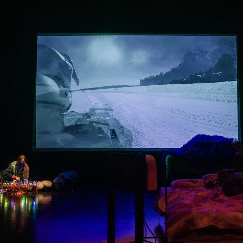 black box performance space with a large projection of a person riding a motor sledge in a wintery landscape on the background. A person is DJing on the floor in front of the projection. The setting is dim but filled with colourful fairylights