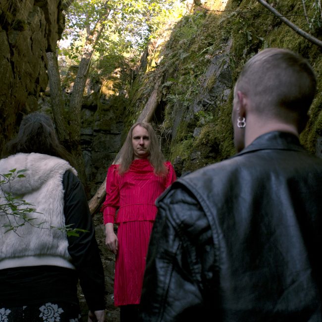 In the photo there are three humans standing in a forest in the middle of moss covered rocks. Two of them are facing away from the camera while one in the middle is looking straight at the camera intensly. The middle one is wearing a red colored outfit while the other two are wearing mostly black but also white.