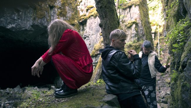 Kolme ihmistä seisoo metsässä kallioiden välissä. Puna-asuinen henkilö istuu kyykkyasennossa kiven päällä katse suunnattuna alas päin. Toinen taas nojaa kivenlohkareeseen ja katsoo sivulle. Kolmas seisoo taaempana kuvassa ja katsoo alas päin.