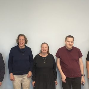 Five people facing the camera stand against a white wall. They smile.