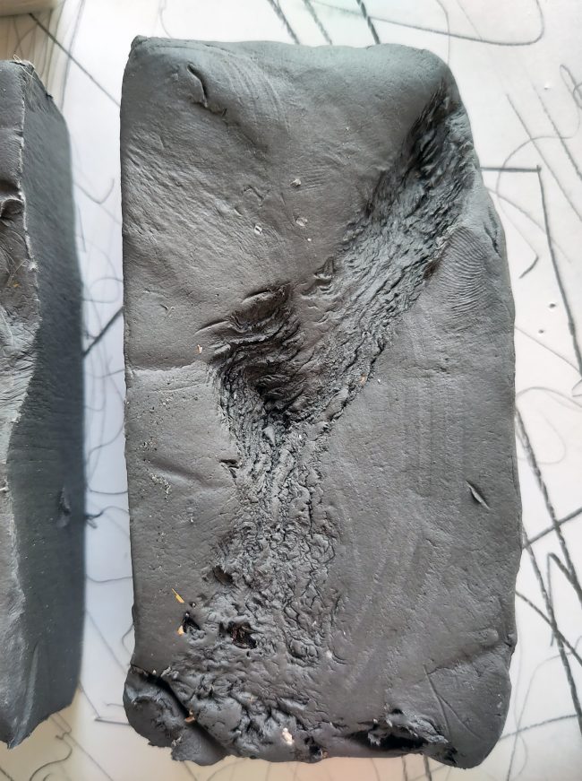 The soft clay mass has been pressed with the roots of a water plant, creating an imprint on its surface.