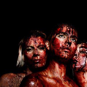 In a portrait composition, there are three blood-covered individuals who appear to be female. The individuals at the edges lean towards the person in the center. The gaze of the person in the center is upward. One of the individuals at the edge looks downward, and the other one looks toward the camera. Their expressions are calm, even melancholic. The background is completely black.