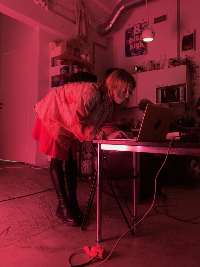 Kuvassa DJ Keiska on kumartunut pöydällä olevan tietokoneen ylle. Kuvassa vallitsee tunnelmallinen punainen valaistus.