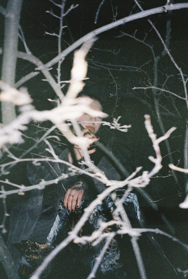 DJ MORA seisoo puun takana, jonka oksat peittävät suurimman osan hänen kasvoistaan ja vartalostaan. Ulkona on pimeää, ja kuva on otettu salamalla.