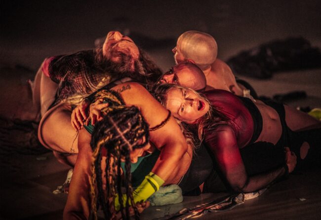 Six people lay in a pile. One's face is visible. Their mouth and eyes are open. The light is warm.