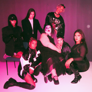 In a slightly hazy, club-like image, seven members of Sara Melleri's team are posed against a magenta-colored background. Four people are sitting or squatting in the front row, and behind them are three others, one of whom is also seated.