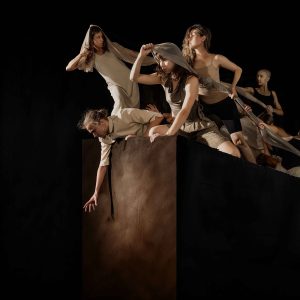Six performers in earthy neutral tones are posed mid-performance on a tall, brown rectangular platform against a black background.